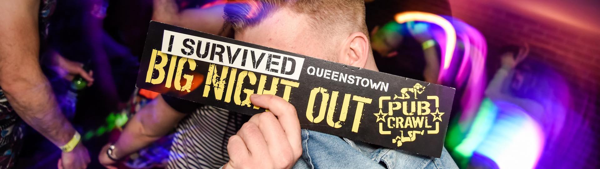 Lad holding I survived the Big Night Out Pub Crawl Queenstown sign with glow stick lights behind him